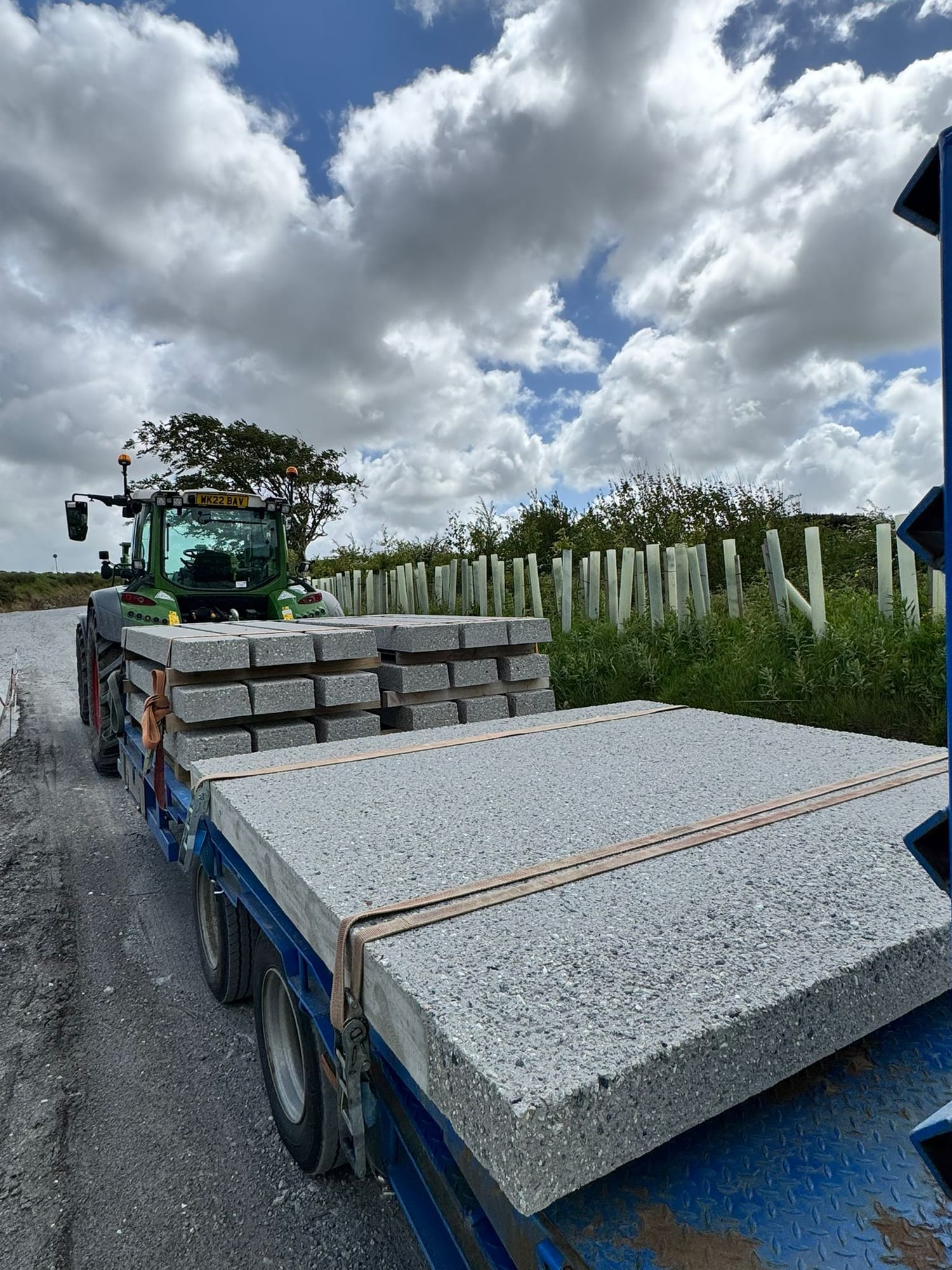 Precast concrete lintels and slabs loaded on a blue flatbed with tractor, leaving the WCCP yard at Chilsworthy, Devon with fence posts visible behind