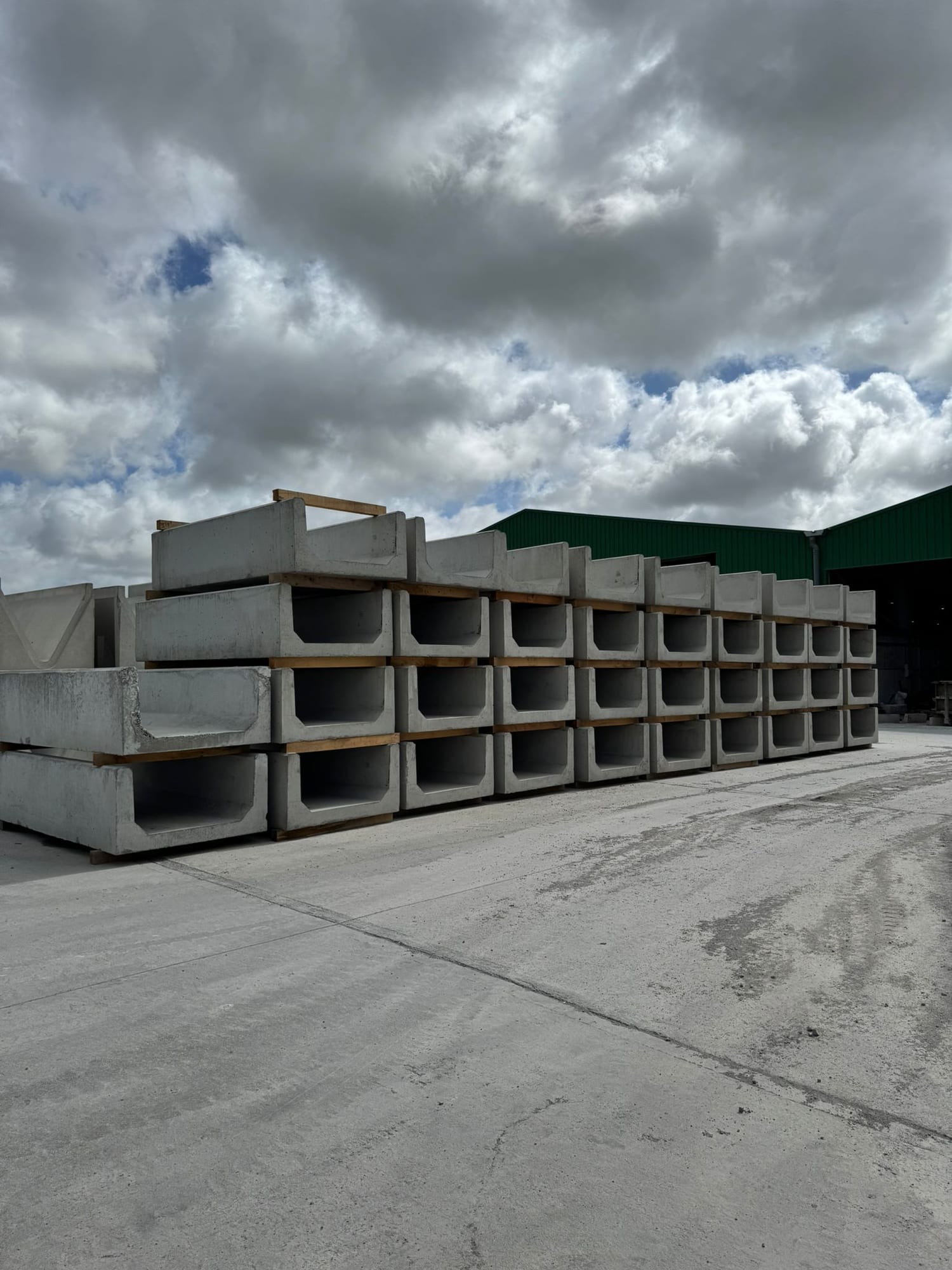 Long rows of precast concrete U-channel slurry sections stacked four-high at the WCCP yard with green-roofed factory building behind, Chilsworthy, Devon